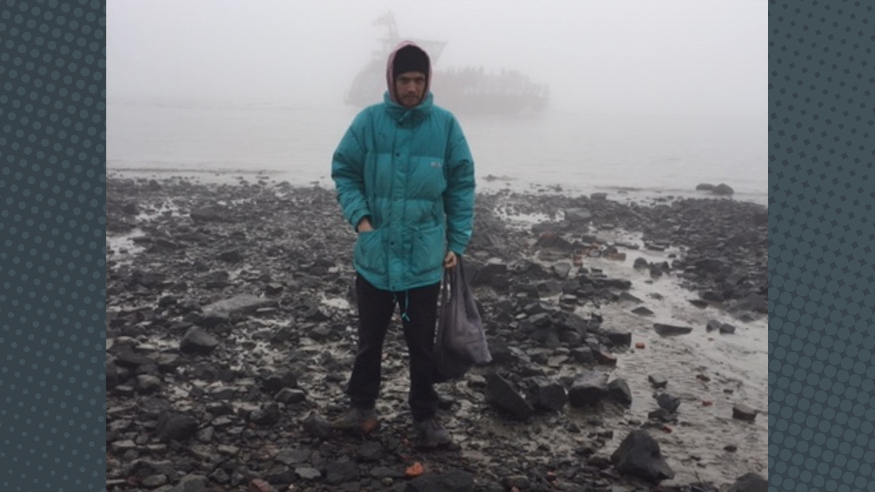 Der Künstler Nutia steht mit Mütze und Regenjacke an einem sehr nebeligen Küstenabschnitt, im Hintergrund sieht man einen Frachter auf dem Wasser.